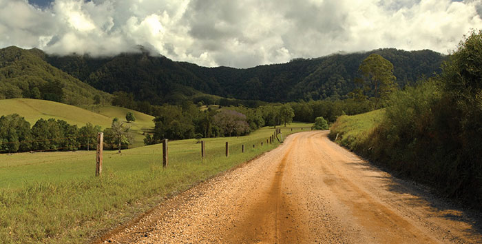 dirt road through paddock with dark tree covered mountains in background with cloudy grey sky