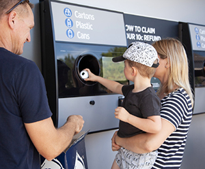 a woman lifts a young boy up to place a can in a return and earn machine while
