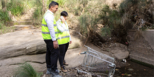 two officers in hiviz jackets inspecting a dumped shopping trolley in a creek