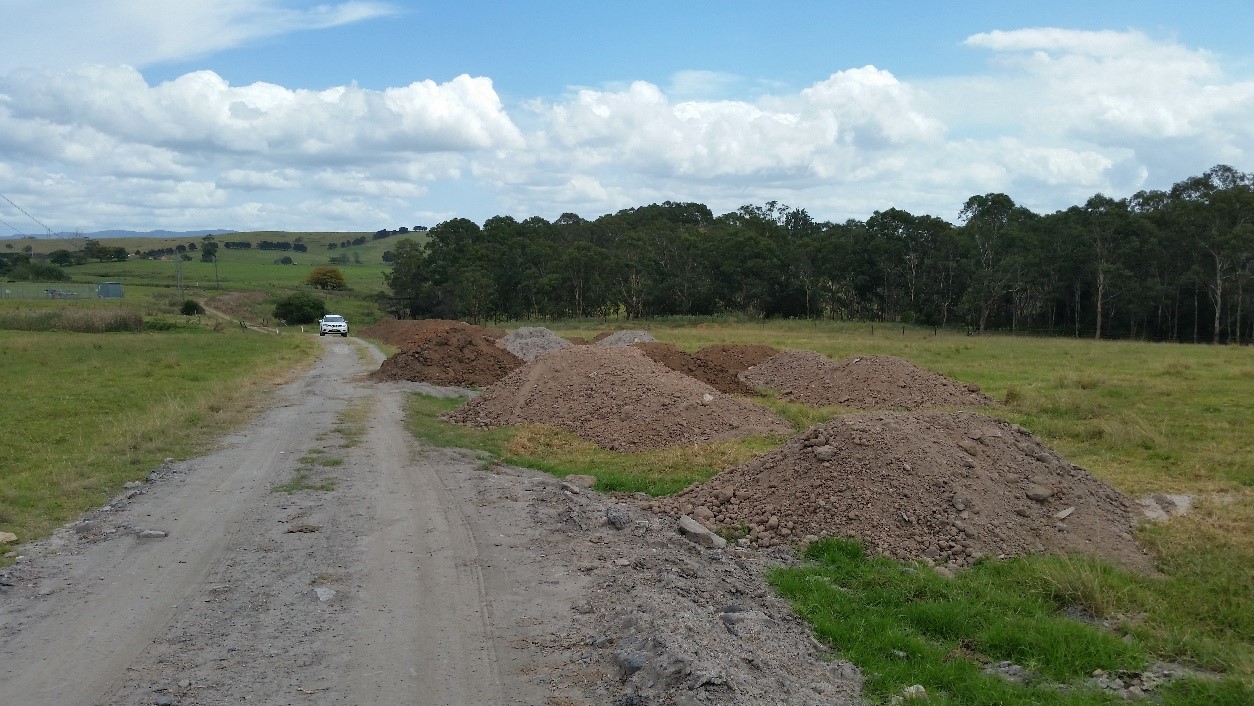 Several piles of contaminated fill at the Croom site sit next to a dirt round which runs through a grass field with trees in the background.